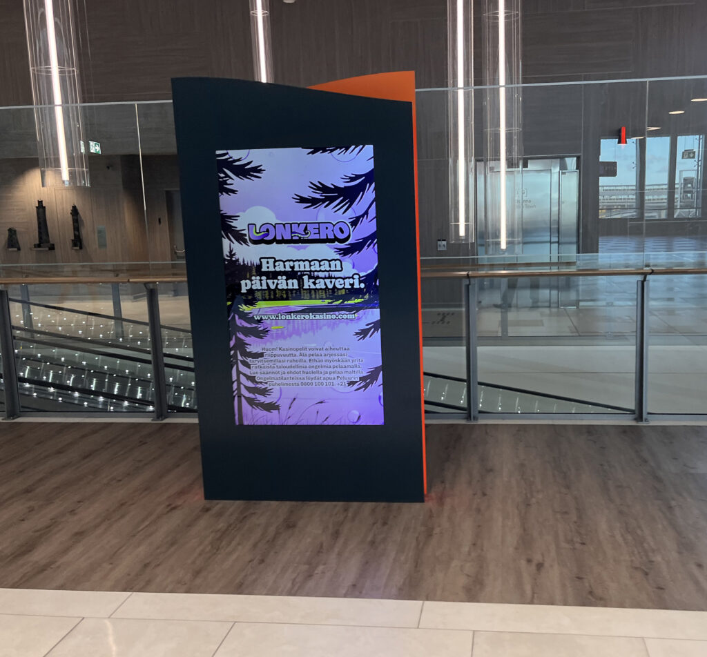 Lonkero Casino mainos kertoo olevansa "Harmaan päivän kaveri." Kuva Tallinnan satamasta, modernista A-Terminaalista (Tallink). Toinen kerros, taustalla loivat liukuportaat alas. 
