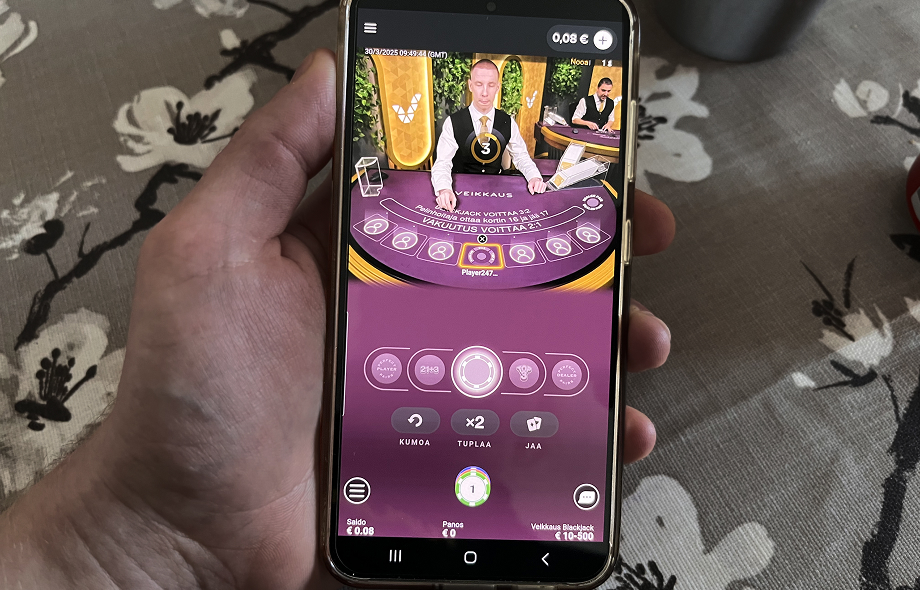Älypuhelin näyttää Veikkauksen live-blackjack-pöydän suomalaisella jakajalla.