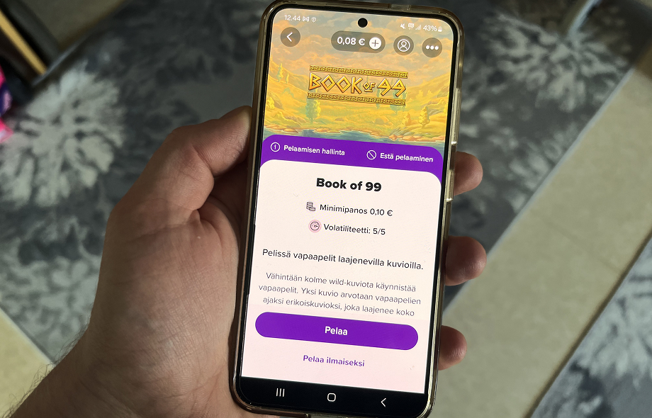 Puhelimen näytöllä Book of 99-kolikkopelin esittely, jossa näkyvät minipanos ja volatiliteetti.