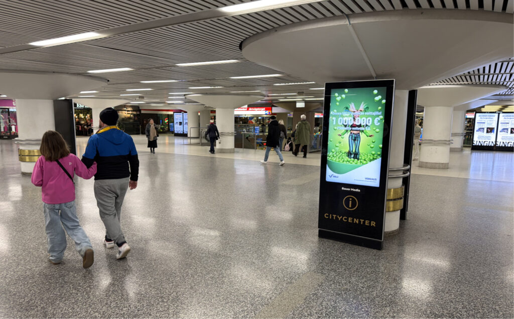 Milli-pelin mainos Helsingin rautatieasemalla onnittelee Milli porukan voittajia. Ruudussa isolla 1 000 000 € ja taustalla Millin arvontapalloissa hyppäävä nainen. Alapuolella Veikkaus logo. Mainoskylttiä operoi Bauer Media ja siinä näkyy myös Citycenter-logo. Mainoksen ympärillä kävelee ihmisiä. Mainos on Citycenterin puolella metroaseman lähettyvillä. 