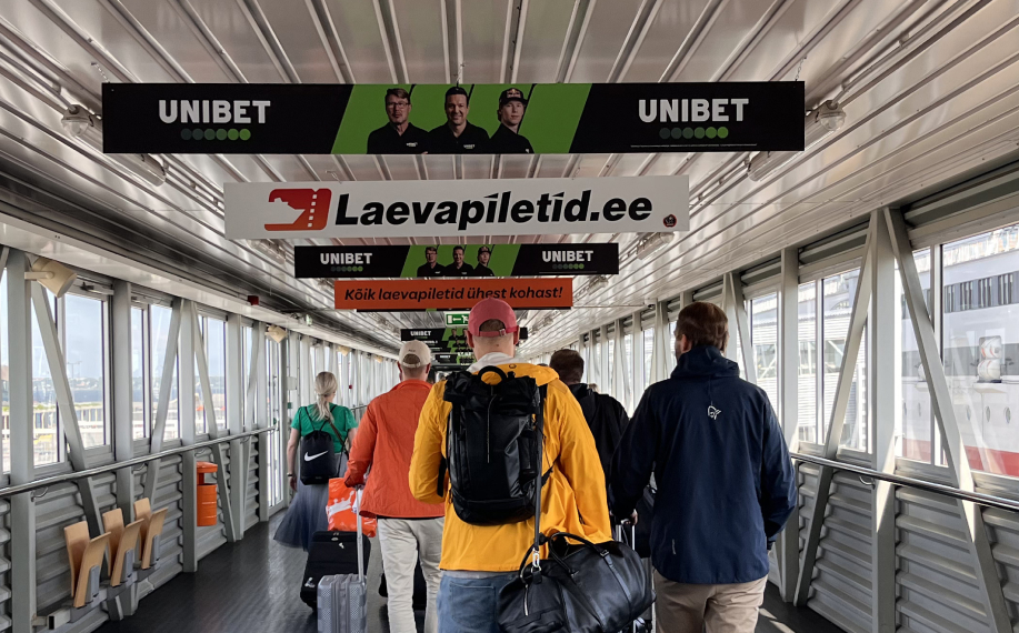 Kuvassa laivamatkustajia satamassa, ikkunasta näkyy Viking Line Xpress. Kattoon asetettu mainoksia kuten Unibetin kylttejä missä näkyy Mika Häkkinen, Jari Litmanen ja Kalle Rovanperä.