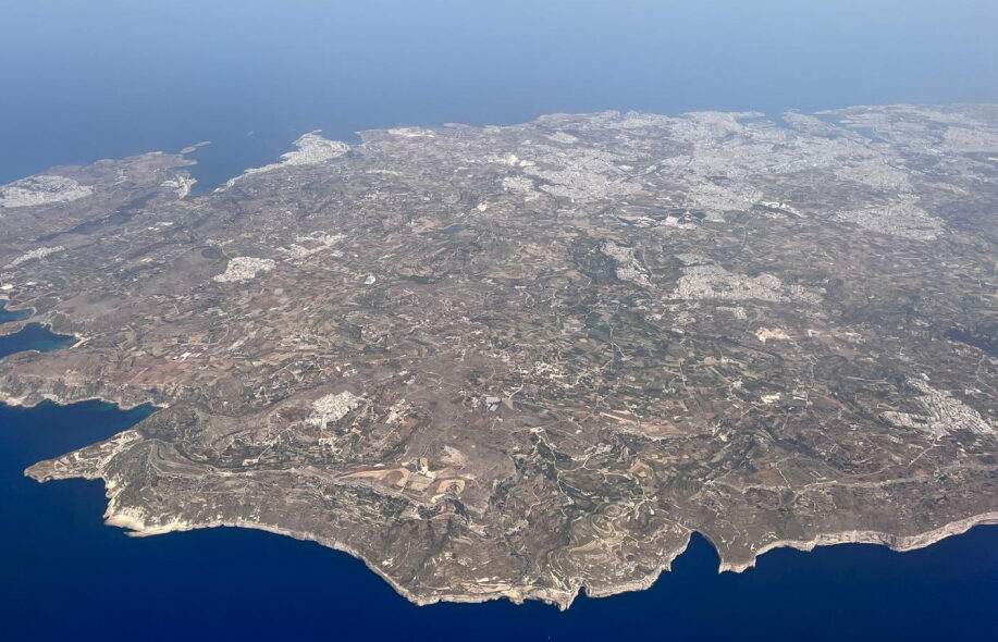 Maltan saari lentokoneesta kuvattuna. Pääkaupunki Valletta ja Grand Harbor näkyvät yläkulmassa. Kallioista etelärannikkoa etualalla.