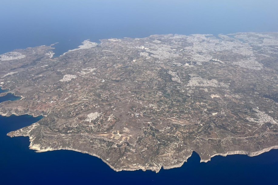 Maltan saari lentokoneesta kuvattuna. Pääkaupunki Valletta ja Grand Harbor näkyvät yläkulmassa. Kallioista ja peltoista etelärannikkoa etualalla. 