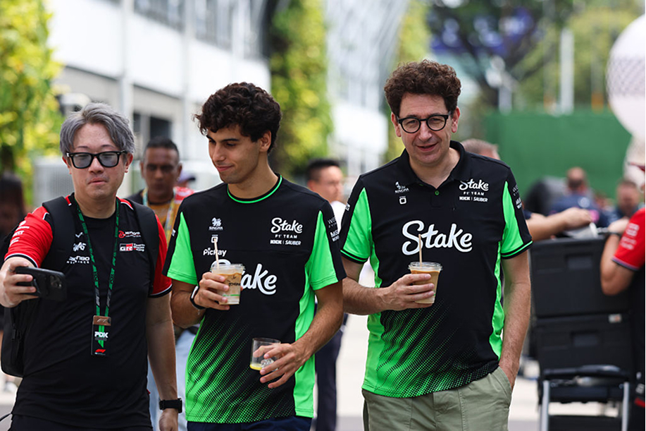Mattia Binotto ja Gabriel Bortoleto ottavat kuvaa fanin kanssa kävellessään kahvit kädessä. Stake F1 tiimin jäsenillä suuri Stake logo paidassaan.