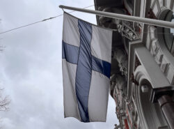 Suomen lippu tummanpuhuvalla taivastaustalla koreaa helsinkiläistä Jugend-rakennusta vasten.