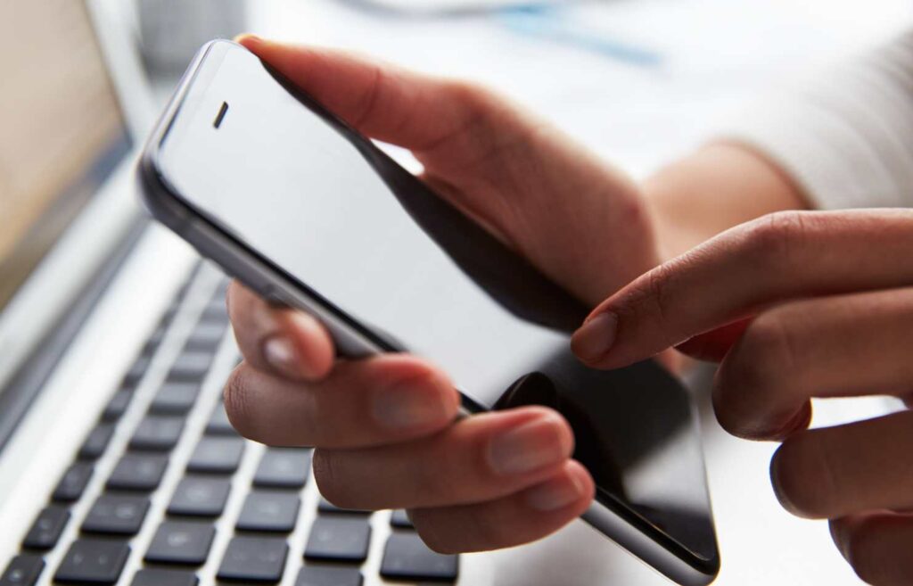 Kuvassa henkilön kädet ja mobiililaite, taustalla kannettava tietokone