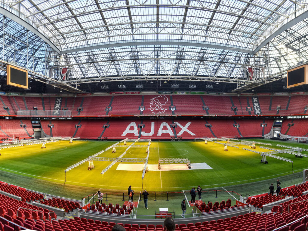 Lasikattoinen hjalkapallostadion. Punaiset penkit ja niissä näkyy isolla AJAX-joukkueen logo. Stadionilla on käynnissä muutostoimintoja koska kentällä näkyy strusseja.