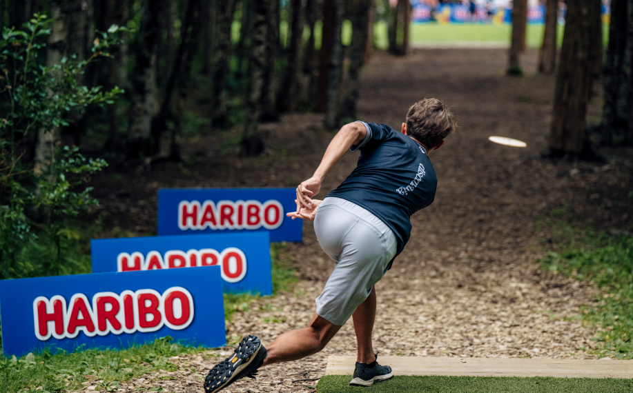 Mies heittää frisbeetä urheilullisesti puiden välikön läpi. Vieressä Haribo-mainoksia. Taustalla puiden takana näkyy yleisöä.