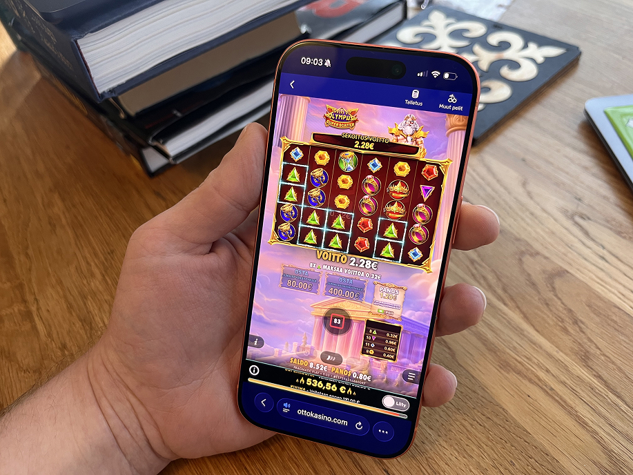Henkilö pitelee älypuhelinta kädessään ja pelaa Otto Kasinolla Pragmatic Playn Gates of Olympus -kolikkopeliä, näytöllä näkyy 2,28 euron voitto ja panostiedot.