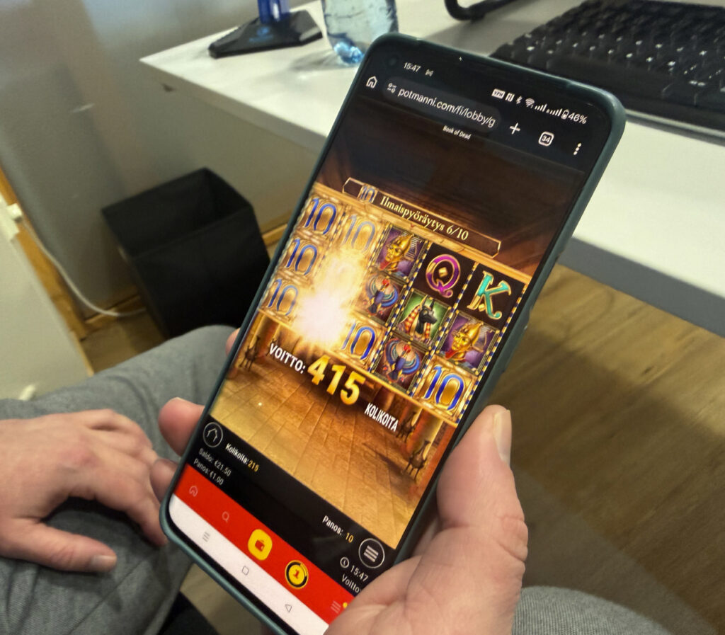 Henkilö pelaa Potmanni-kasinolla Book of Dead -kolikkopeliä mobiililaitteella, näytöllä näkyy 415 kolikon voitto ja ilmaiskierrosten eteneminen.