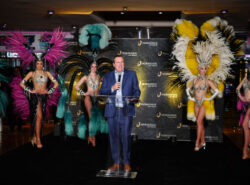 Tom Reeg pitää puhetta kasinon avajaissa. Hänen takanaan on karnevaaliteemaisesti pukeutuneita naisia.