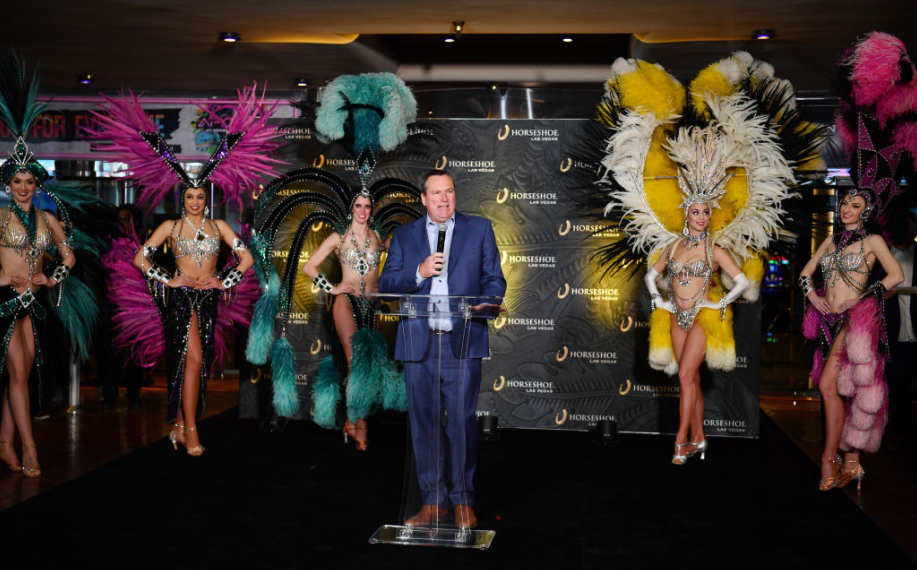 Tom Reeg pitää puhetta kasinon avajaissa. Hänen takanaan on karnevaaliteemaisesti pukeutuneita naisia.