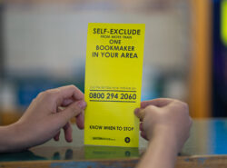 Henkilö pitää käsissään kirkkaankeltaista esitettä, jossa kerrotaan rahapelaamisen itse-estosta ja annetaan maksuton brittiläinen tukipuhelinnumero.