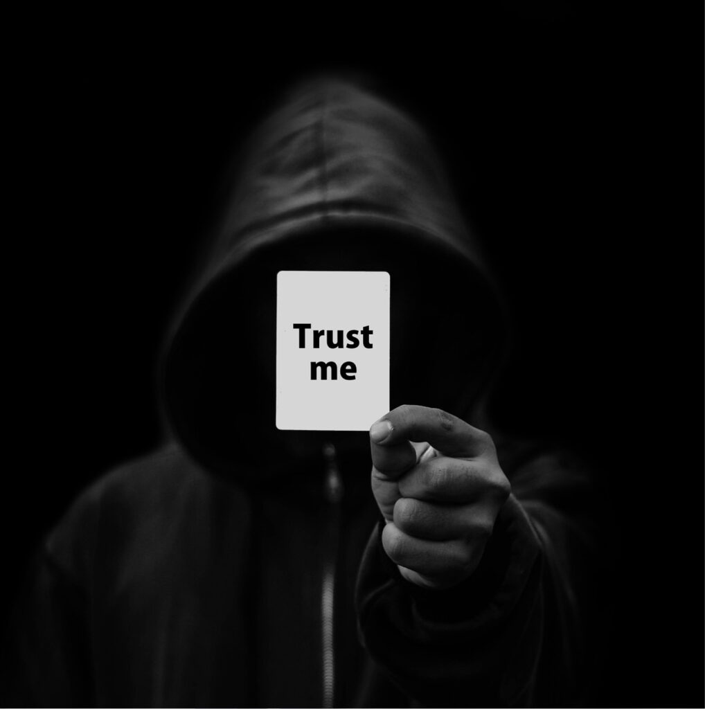 Huppupäinen henkilö pitelee “Trust me” -korttia pimeässä, kuvaten internetin anonyymiä ja harhaanjohtavaa tietoa.