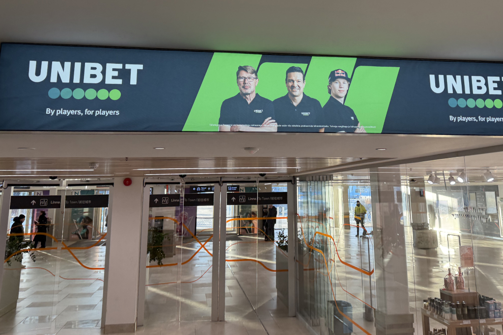 Unibet mainos Tallinnan satamassa  ja vihreällä brändibannerilla uloskäynnin yläpuolella. Mainoskasvoina Mika Häkkinen, Jari Litmanen ja Kalle Rovanperä, joilla kaikilla on päällään Unibet-paita. Rovanperällä on myös RedBullin lippis.