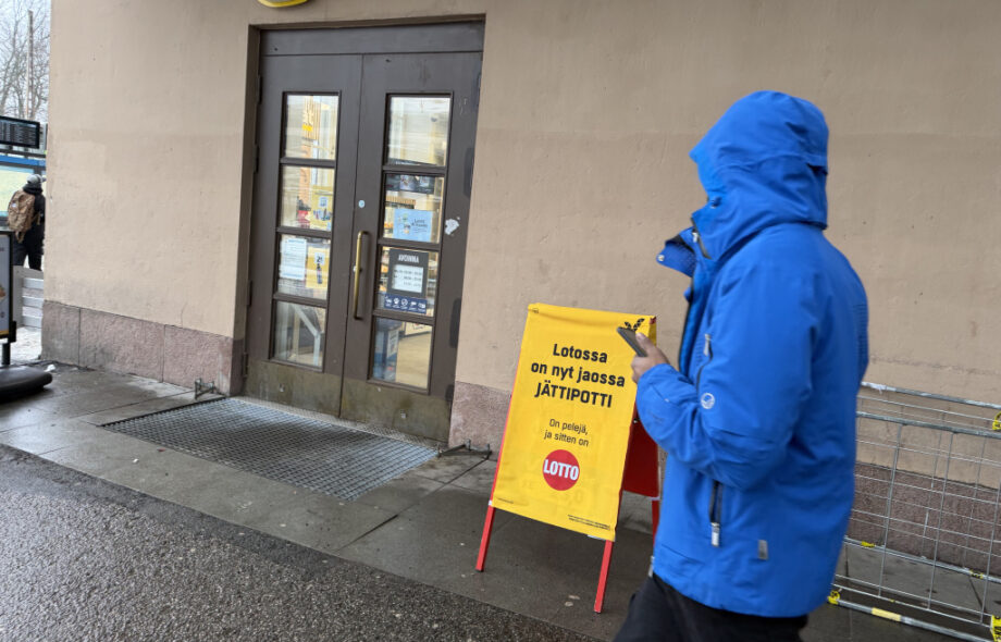 Veikkaus Lotto-mainos Helsingin Rautatiaseman ulkopuolella märässä ja synkässä kelissä.