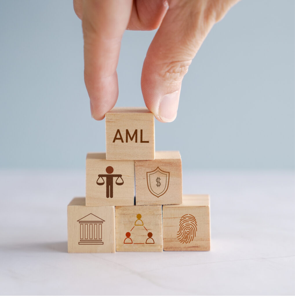 Käsi asettaa pinoon rahanpesun estämistä kuvastavia puupaloikoita. Esimerkiksi palikka missä lukee "AML " tai toisessa palikassa sormenjälki.