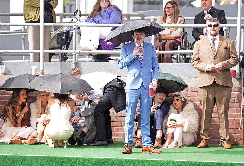 Katsojia Aintreen Grand National -laukkakilpailussa sateessa, osa yleisöstä sateenvarjojen alla, etualalla pukuihin pukeutuneita katsojia juomien kanssa katsomon edessä.