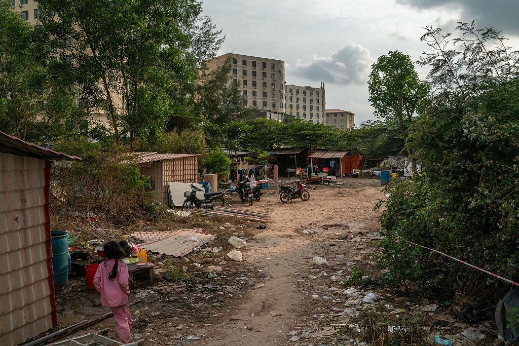 Roskaantunut ja karu piha-alue Sihanoukvillessa Kambodžassa. Etualalla lapsi kävelee hökkeleiden ohi, taustalla näkyy korkeita rakennuksia ja kasvillisuutta.