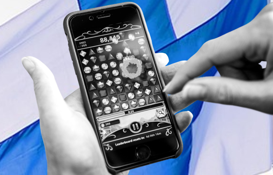 Henkilö pelaa mobiilipeliä älypuhelimella Suomen lipun edessä.