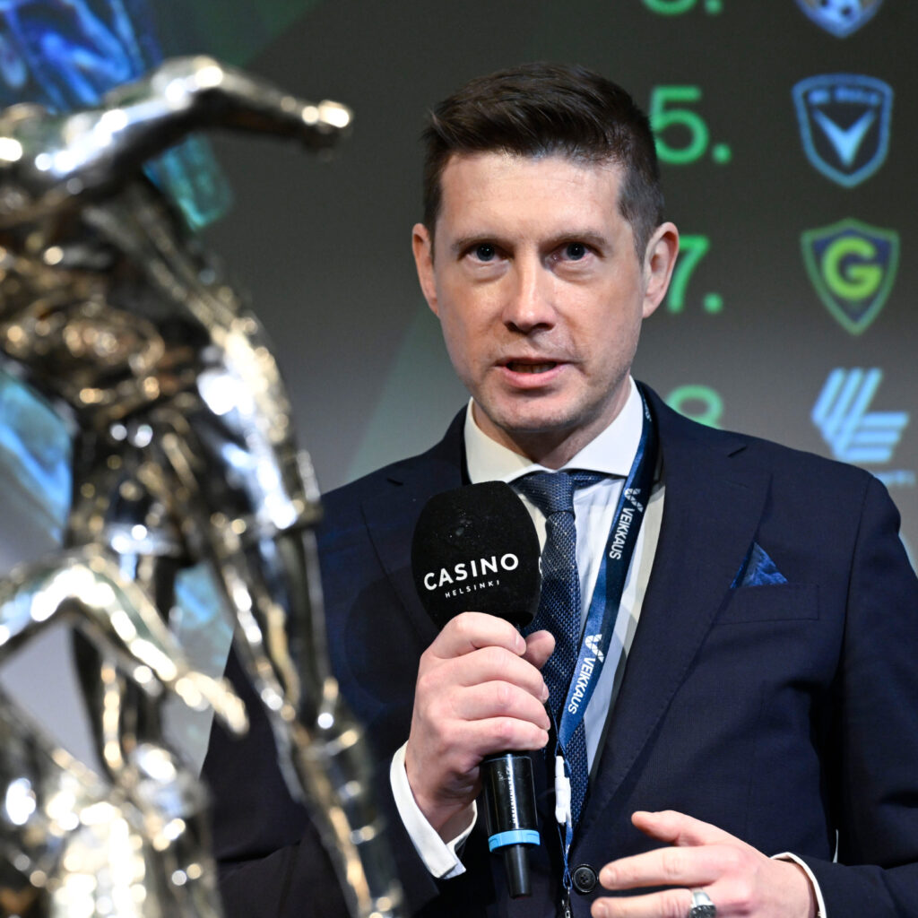 Pukuun pukeutunut Timo Marjamaa pitää kädessään Casino Helsinki mikrophonia ja kaulassaa Veikkaus-logollinen avainketju. Etualalla Veikkausliigan pokaali.
