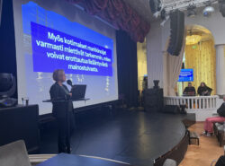 Anna-Riikka Hovi-Taunila esittelee lavalla rahapelimarkkinan markkinointia käsittelevää diaa tapahtumassa, yleisö seuraa salissa.