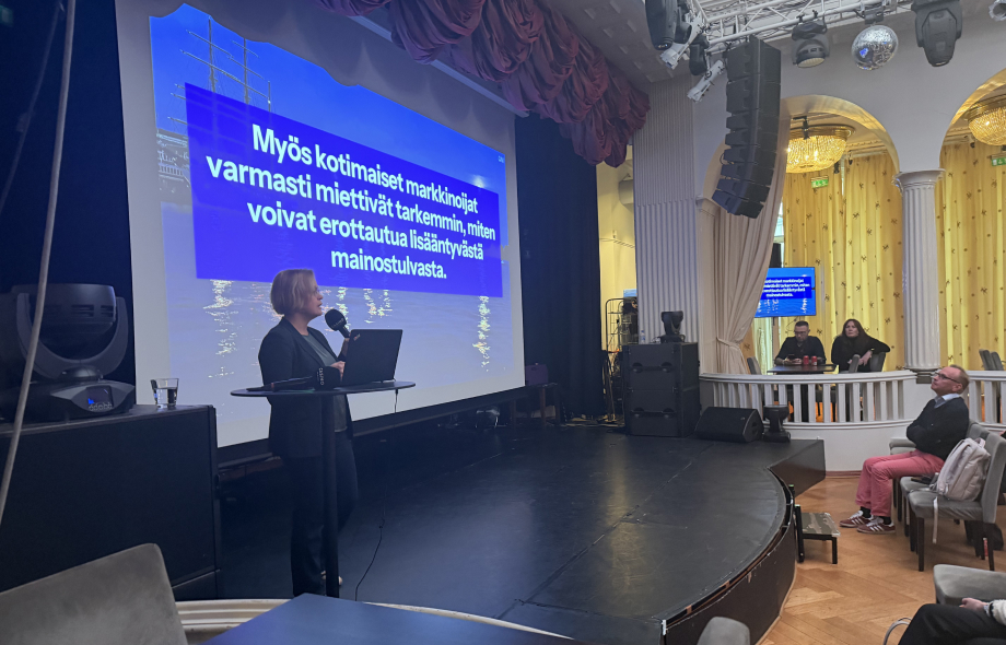Anna-Riikka Hovi-Taunila esittelee lavalla rahapelimarkkinan markkinointia käsittelevää diaa tapahtumassa, yleisö seuraa salissa.