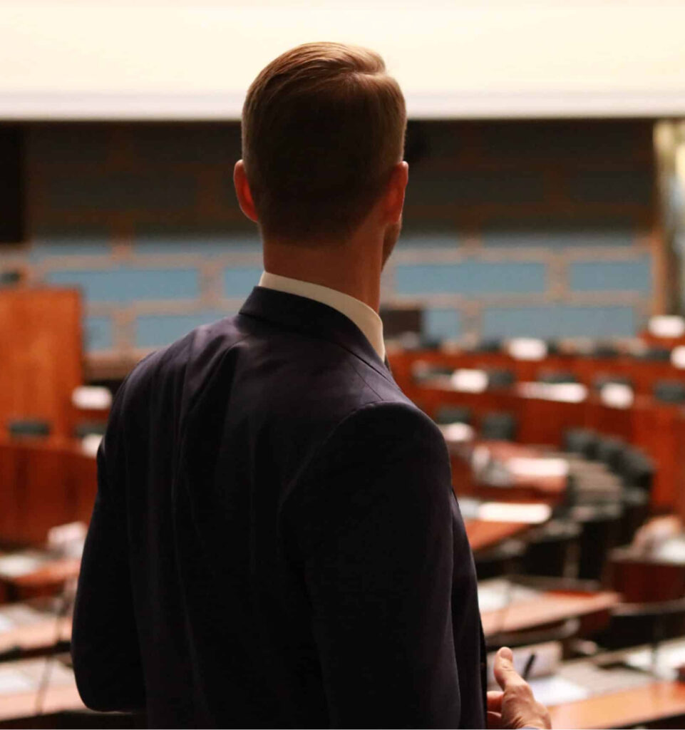 Jouni Ovaska katselee eduskunnan istuntosalia kuvattuna takaapäin.