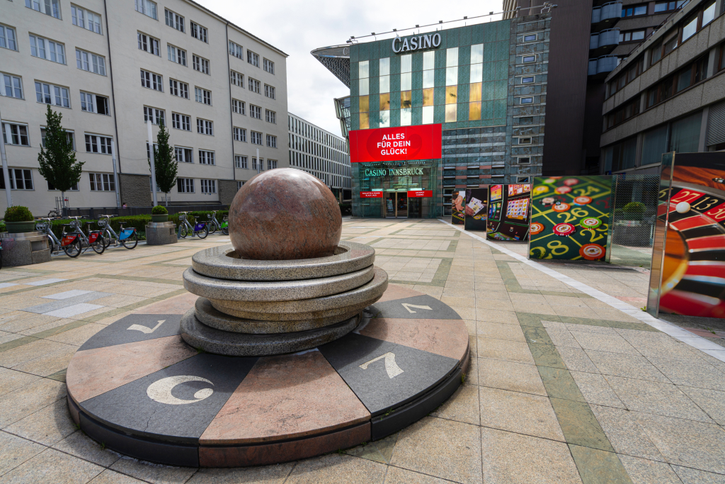 Ruletti-veistos/suihkulähde sekä rahapeliteemaisia mainoskuvia Casino Innsbruckin edessä. Casinon oven yläpuolella olevassa kyltissä lukee "Alles Fur Dein Gluck"