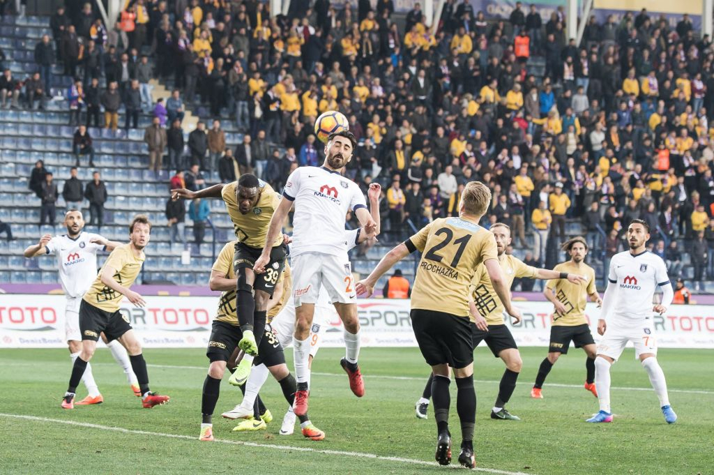 Pelaajia kamppailemassa pallosta kentällä. Taustalla väkilukuisen stadionin katsomon edessä olevilla mainoskylttejä kentälle päin "Sportoto"