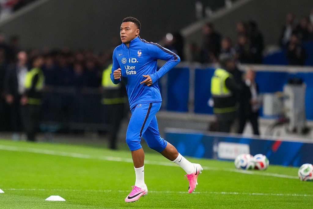 Mbappe juoksee kentälle Ranskan verryttelyasussa. Takissa Volkswagen ID ja Uber Eats logot. Takissa, shortseissa, sukissa ja kengissä näkyy Nike. Taustalla stadion väkeä ja jalkapalloja. 
