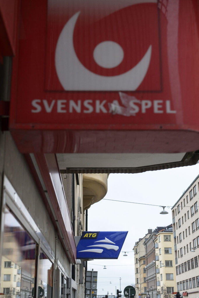 Etualalla Svenska Spel pelaamon katukyltti. Sen takana tarkennettuna ATG kyltti ja hevoslogo. Kaupunkimaisemaa.