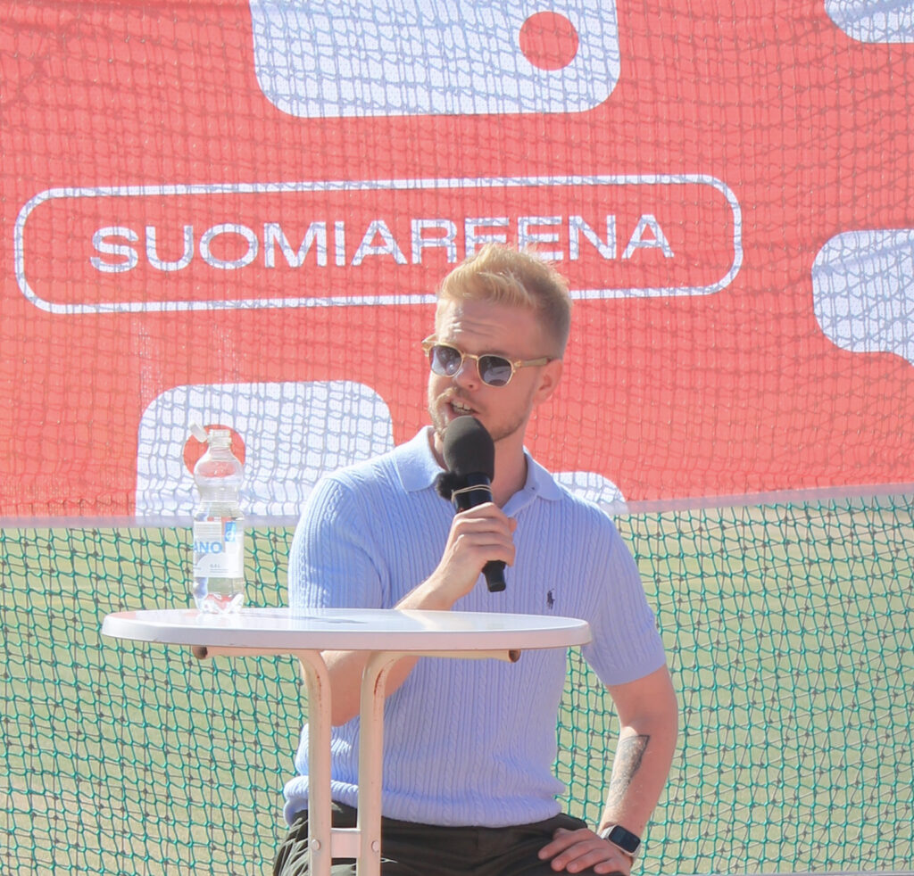 Joel Häkämies kesäisessä asussa kädessään mikrophoni. Taustalla "Suomiareena" logo.
