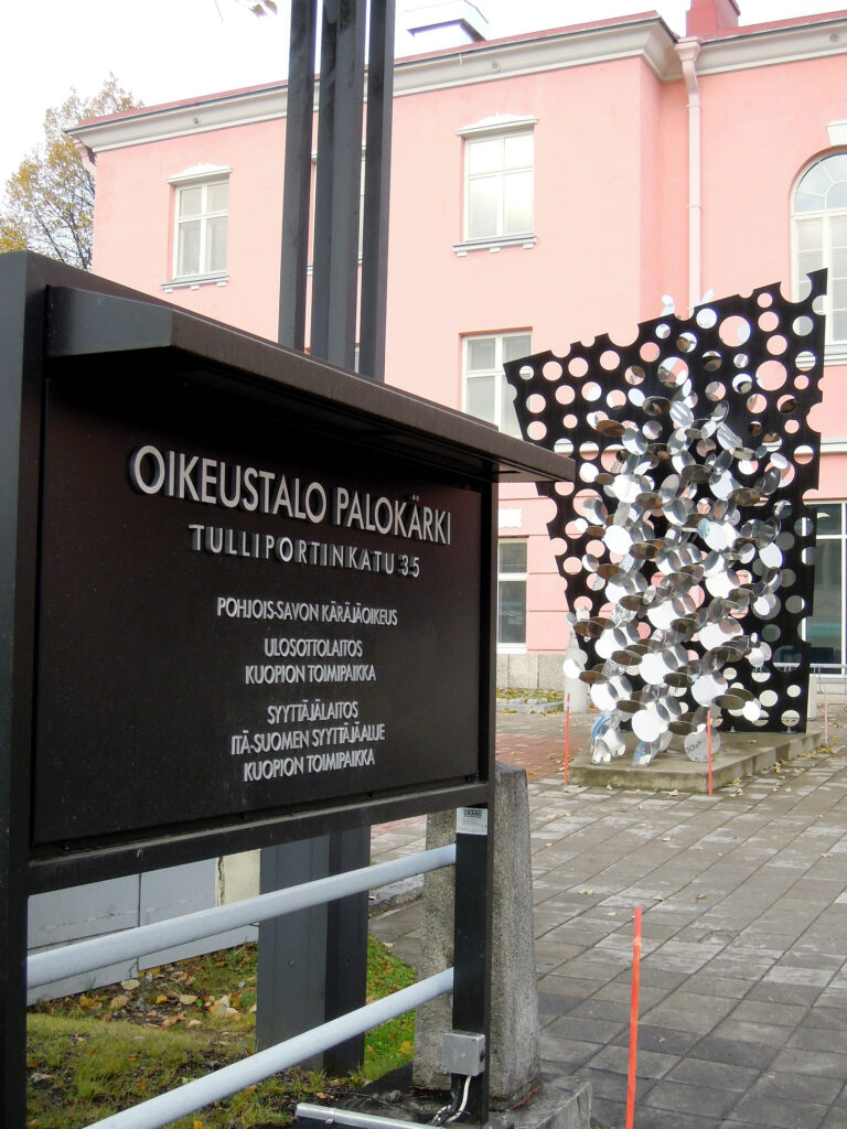 Oikeustalo Palokärki katukyltti. Taustalla rakennus ja metallinen  reikiä täynnä oleva levytaideasennus.