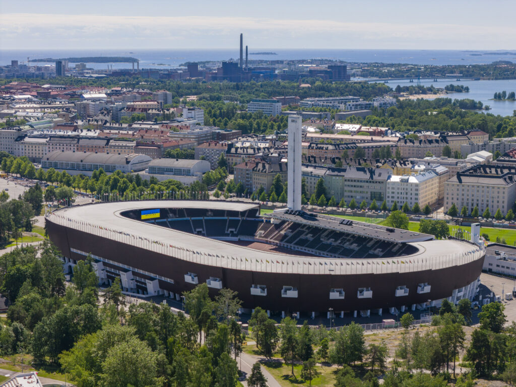 Ilmakuva Olympiastadionista. Stadionin ruudulla näkyy Ukrainan lippu. Taustalla kaupunkinäkymää ja merellistä Helsinkiä, kuten Hietaniemen uimaranta.