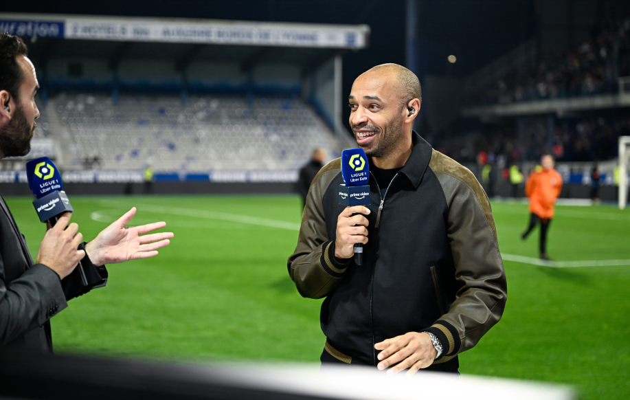 Thierry Henry hymyilee jalkapallostadionilla mikrophoni kädessään. Rento pukeutuminen. Vieressä toinen juontaja.