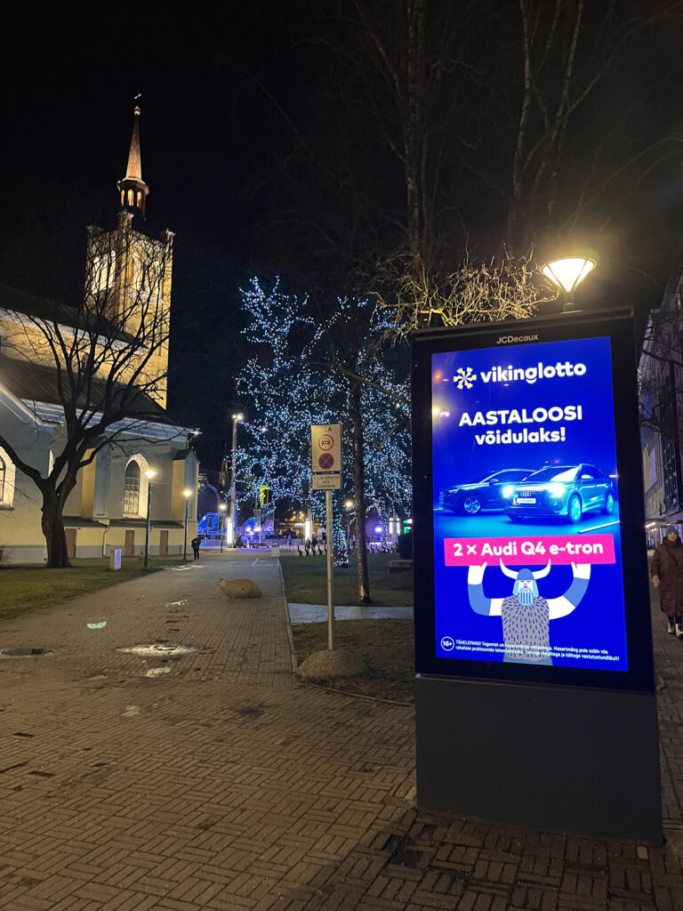 Tallinnan Vabaduse Väljak näkyy taustalla. Etualalla Vikinglotto mainos mikä mainostaa lisäarvonnoista voitettavia Audin autoja. Puissa jouluvaloja. Valaistu kirkko.