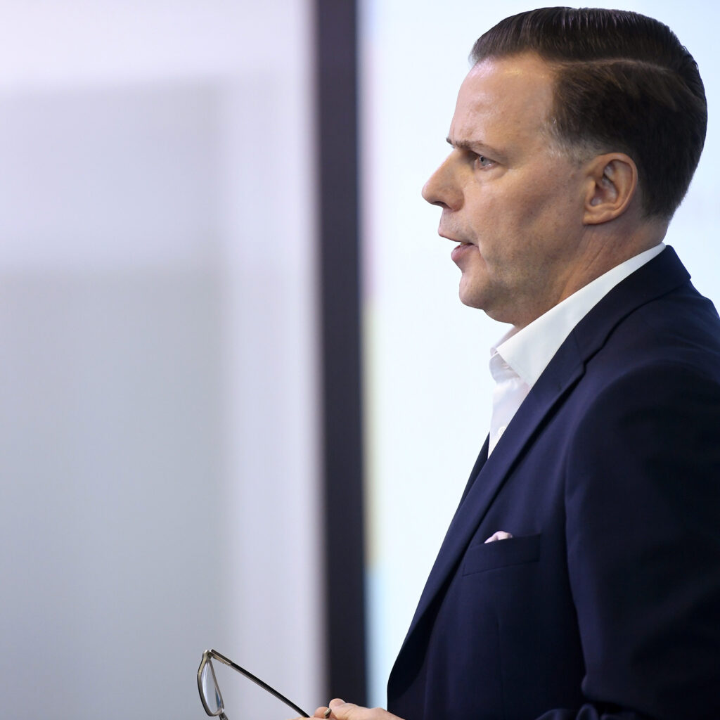Olli Sarekoski. Päällä puku, kädessä silmälasit. Sivunäkymä.