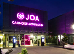 JOA Casino De Montond sisäänkäynti. Moderni tyyli, muutama kasvi näkyy. Liilat ledivalot nimikyltissä.