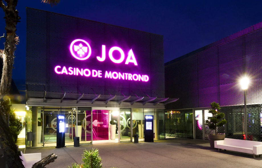 JOA Casino De Montond sisäänkäynti. Moderni tyyli, muutama kasvi näkyy. Liilat ledivalot nimikyltissä.