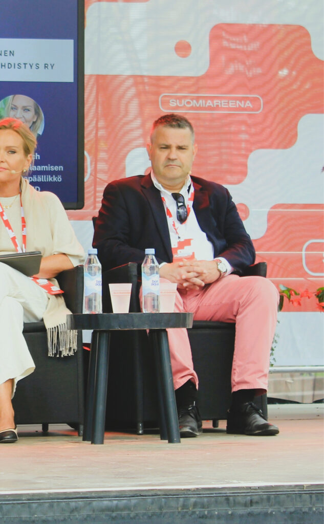 Janne Nikkinen, vieressä näkyy osittain Taina Grach. Panelistit lavalla. Taustalla Suomiareena logo ja väritystä.