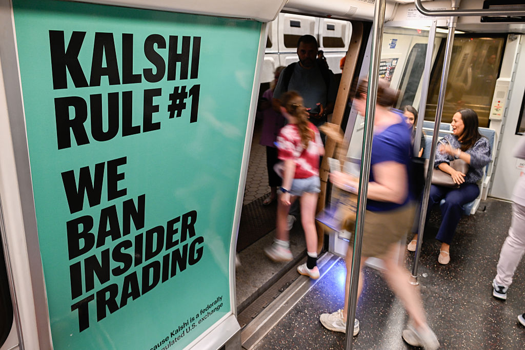 New York metromainoksessa Kalshi mainos: #1 sääntö – me annamme porttikiellon sisäpiirikaupalle. Alhaalla pienellä tekstillä näkee että kalshi on kirjoittanut "Olemme liittovaltion... ...U.S. Exchange". Suuri mainos. Ihmisiä viettämässä elämää metrossa. Lapsi poistumassa laiturille.