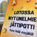 Kadulla ajattomalta näyttävä kyltti kertoo lotossa olevan unelmien jättipotti. Kehottaa kysymään lisää myyjältä.