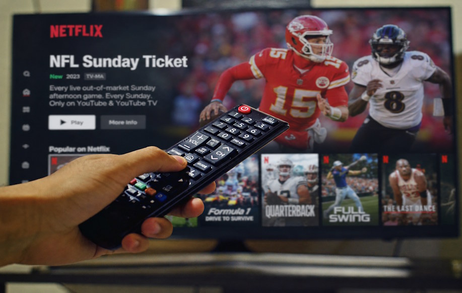 Television ruudulla näkyy Netflixin urheilusisältöä ja NFL-aiheinen ohjelmavalikko, kun katsoja pitää kaukosäädintä kädessään.