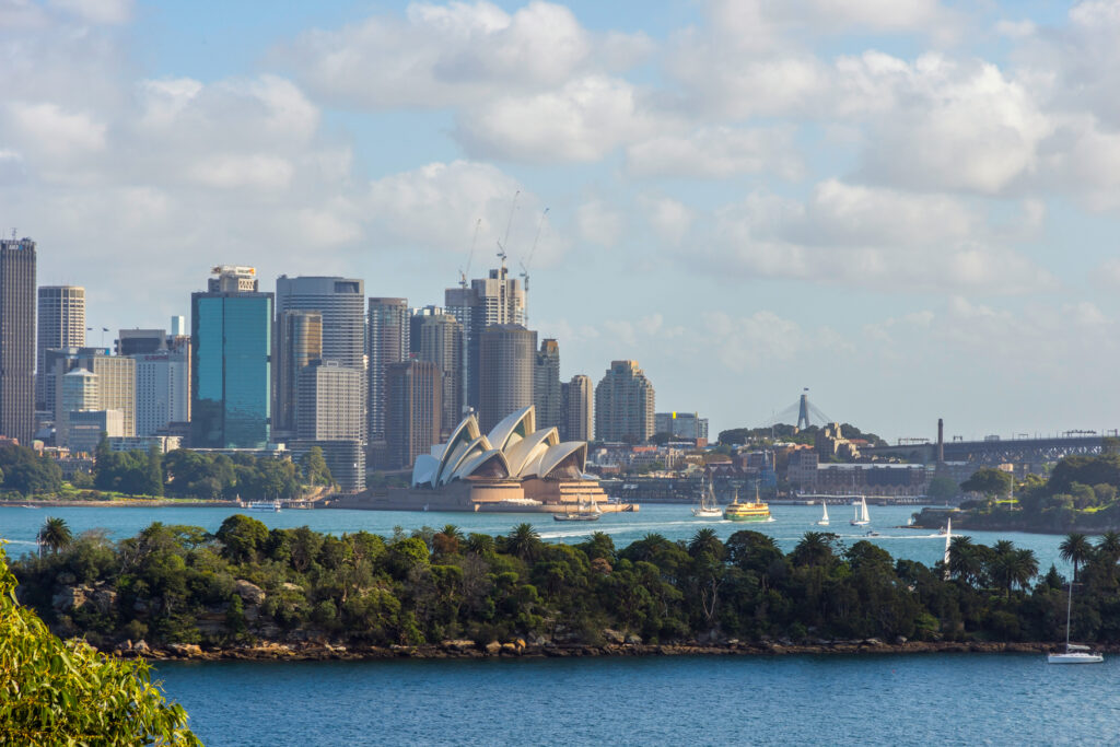 Sydneyn merimaisemaa, etualalla vihreä niemi, taustalla aoopperatalo ja tornitaloja. Suhteellisen kesäinen sää koska paljon veneitä, kuitenkin puolipilvinen sää.