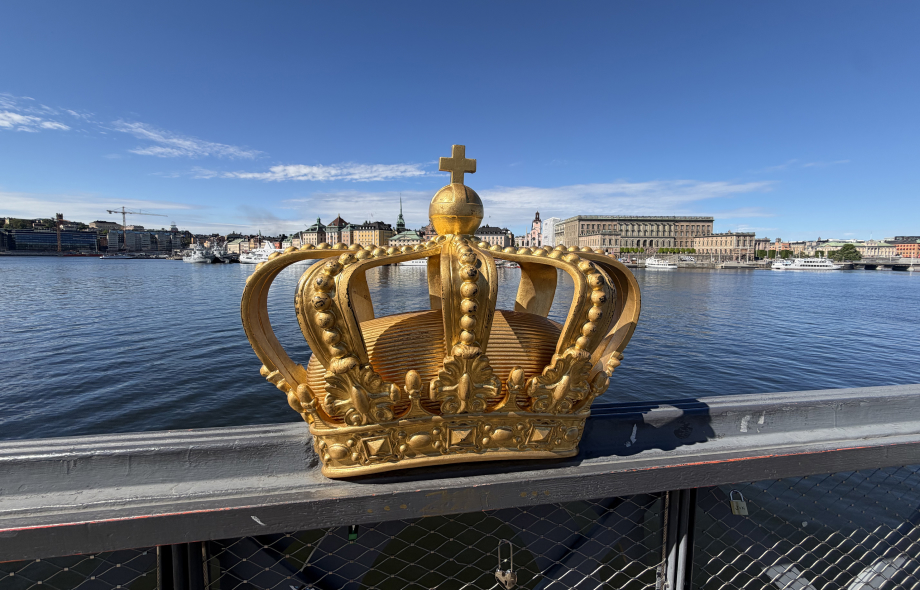 Etualalla sillan kaiteeseen kiinnitetty Kruunu. Taustalla Ruotsin kuninkaanlinna. Kuva toukokuussa 2026.