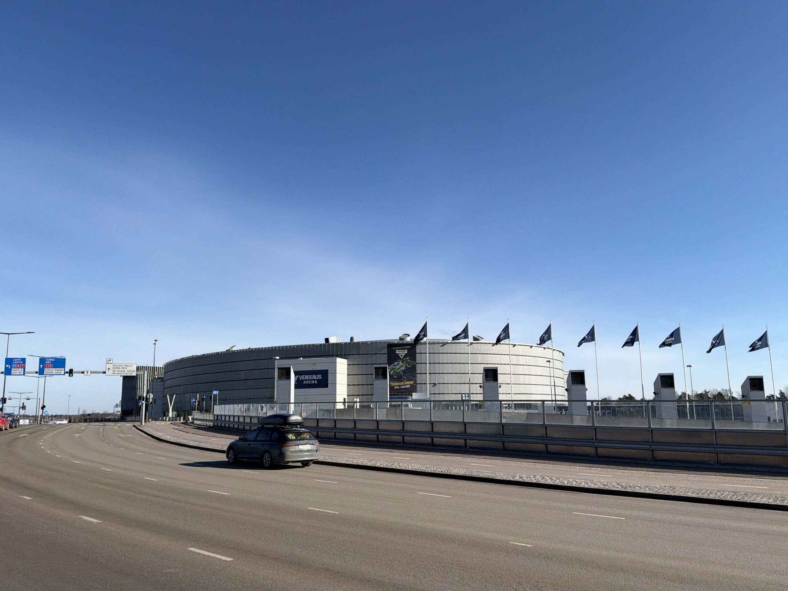 Veikkaus Arena ja sen liput kuvattuna suuren tien takaa. Sininen taivas, ei lunta.
