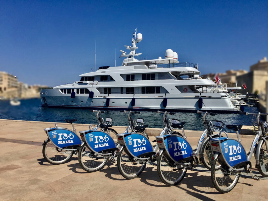 Malta-teemaisia polkupyöriä. Taustalla jahti ja maltalaista rakennettua rantamaisemaa.