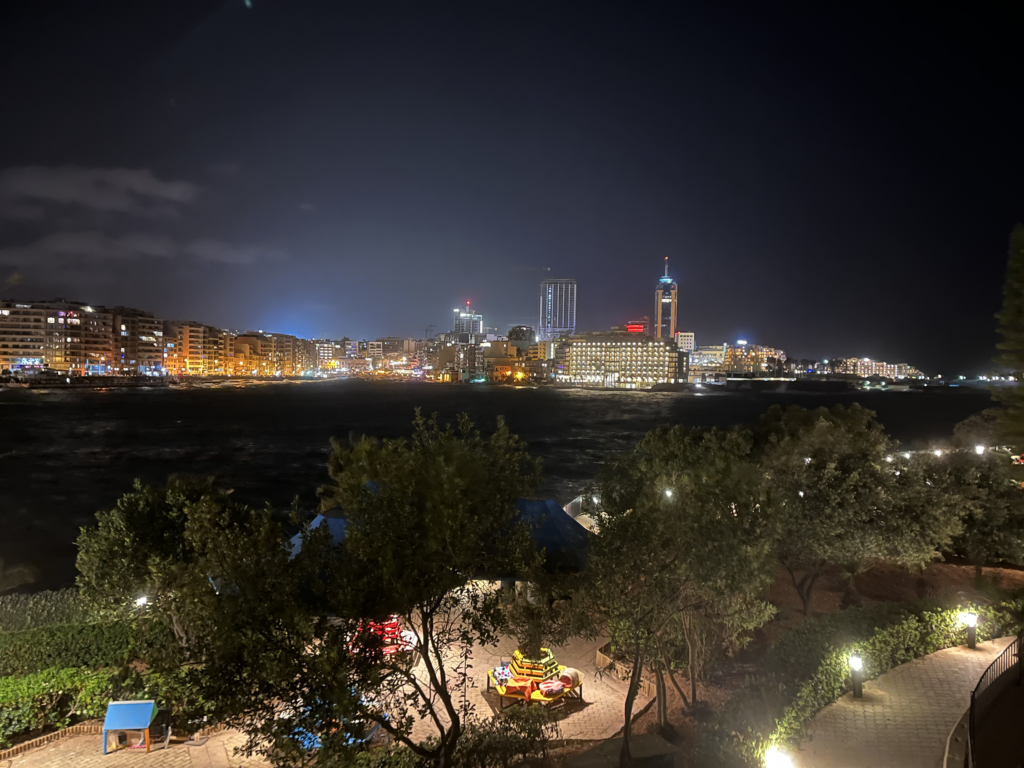 Balluta- ja Spinola-bay iltahämärässä. Paljon rakennuksia. Etualalla puisto. Taka-alalla lahden takana muutama korkea rakennus.