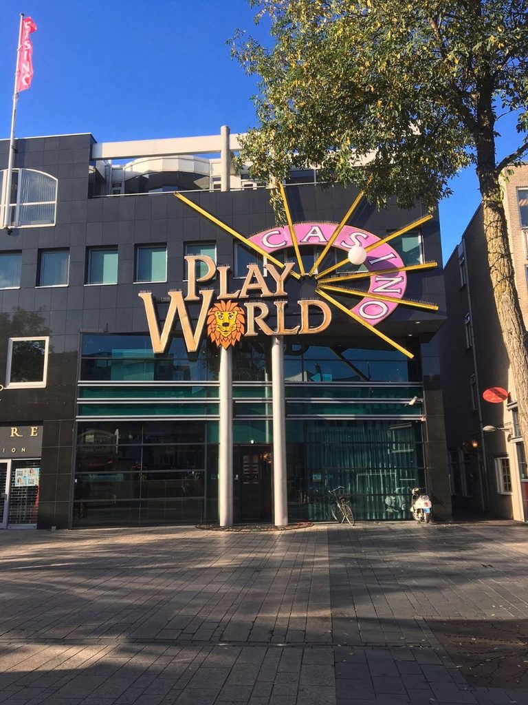 Play World Casino logo rakennuksen seinässä. Etualalla siististi kivitettyä katua ja puu. Muutama fillari parkissa. Sininen taivas. "Casino" lippu.